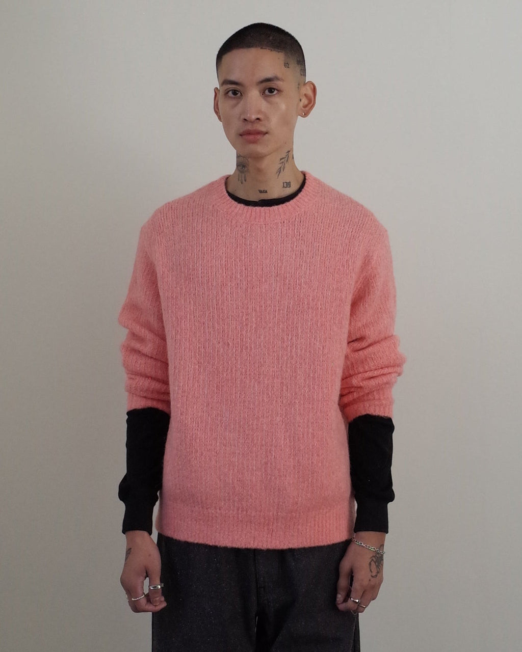 A man stands in a minimalist room with white walls and a dark wood floor, wearing a pink knit sweater over black long sleeves, paired with dark gray trousers and black shoes.