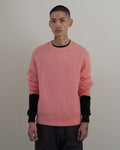 A man stands in a minimalist room with white walls and a dark wood floor, wearing a pink knit sweater over black long sleeves, paired with dark gray trousers and black shoes.