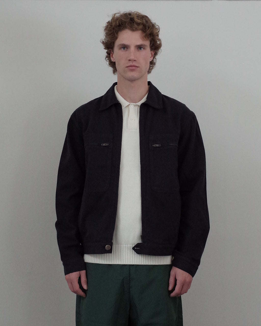 A man with curly light brown hair stands against a light gray wall wearing a dark navy overshirt over a cream knit polo, paired with dark green trousers.