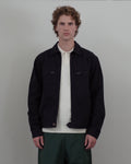A man with curly light brown hair stands against a light gray wall wearing a dark navy overshirt over a cream knit polo, paired with dark green trousers.