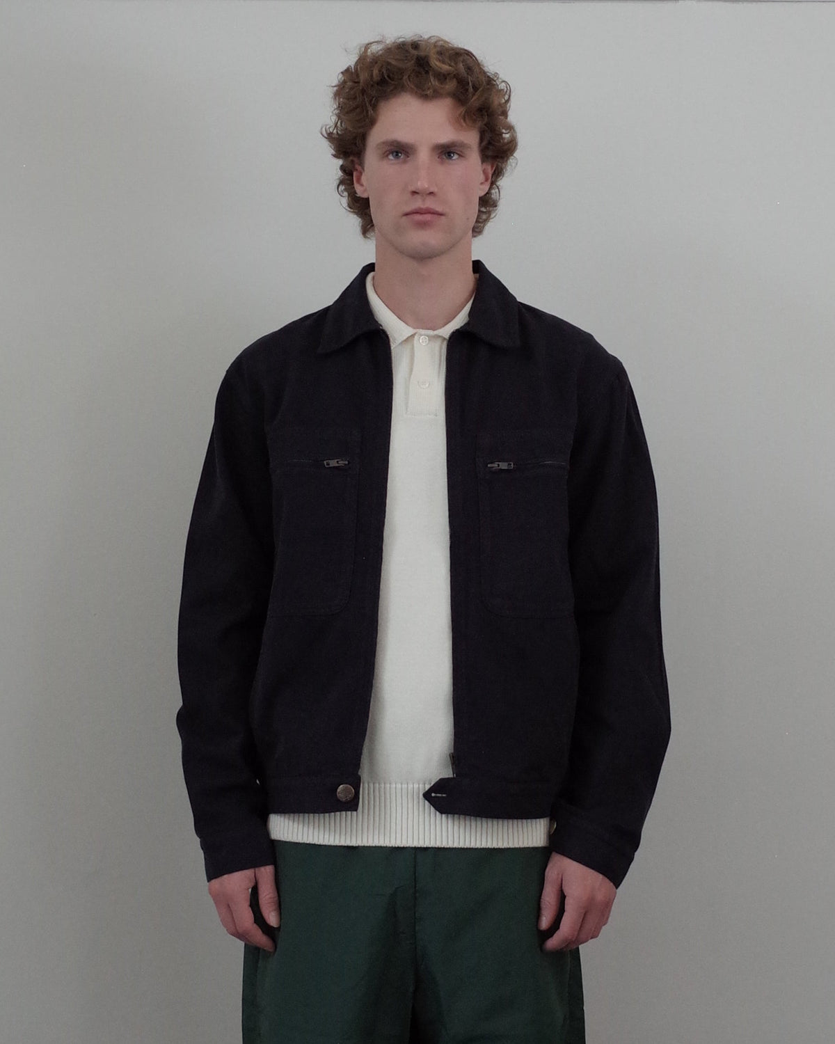 A man with curly light brown hair stands against a light gray wall wearing a dark navy overshirt over a cream knit polo, paired with dark green trousers.