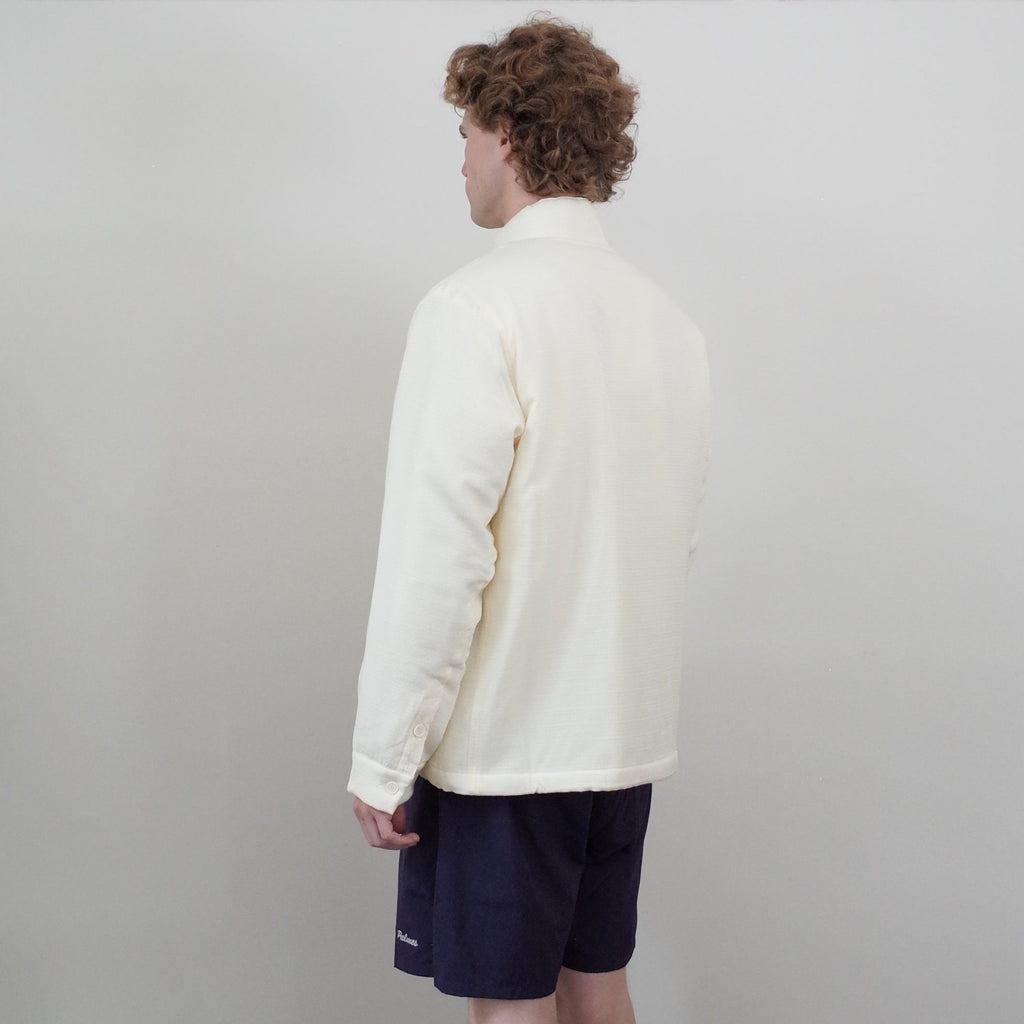 A person standing in a corner of a room with white walls and dark wood flooring, wearing a cream zip-up fleece, navy shorts, black socks, and black shoes, viewed from the back.