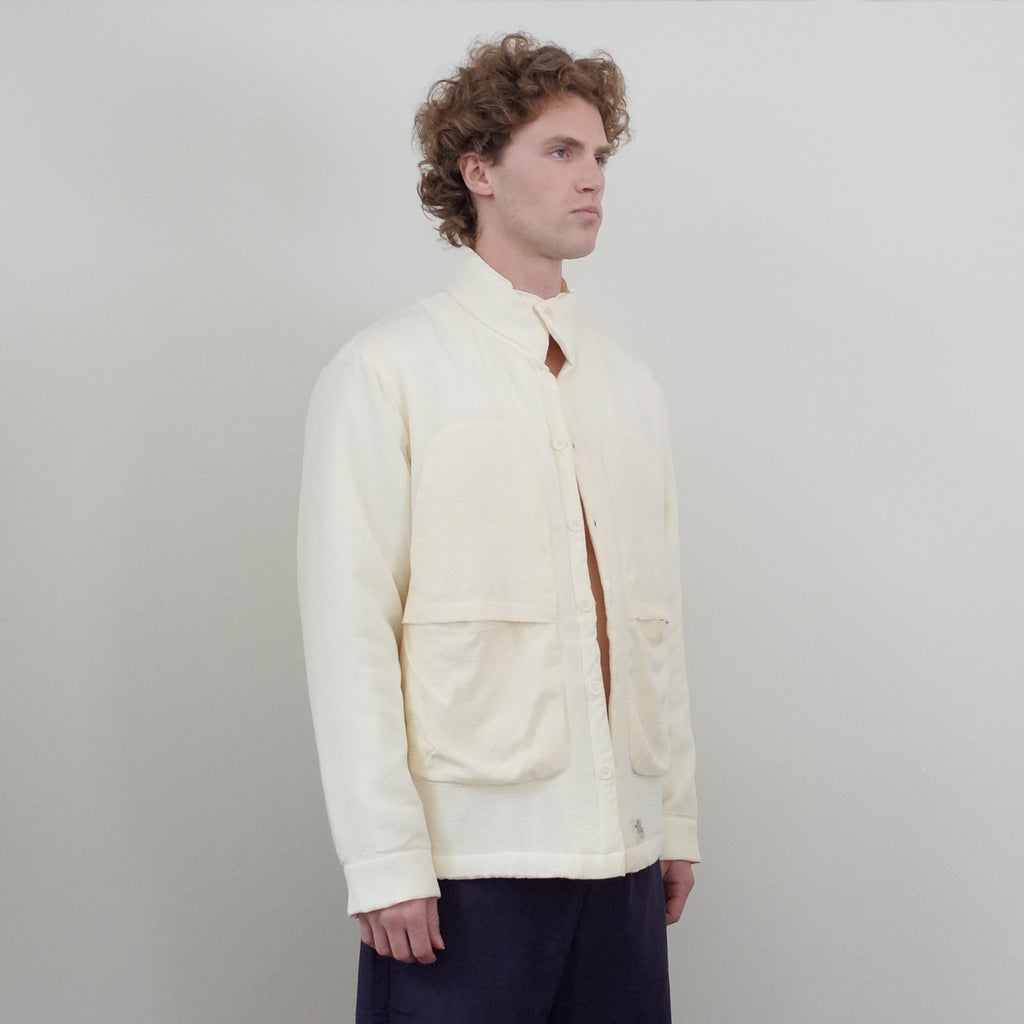 A man stands in a minimalist room with white walls and dark wood floors, wearing a cream-colored zip-up jacket, navy shorts, black socks, and black shoes.