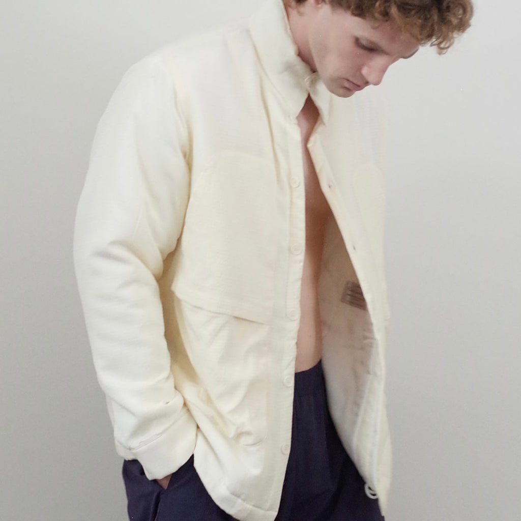 A man wearing a cream-colored, lightweight open-front jacket over a bare torso and dark navy shorts, standing against a plain light gray background.