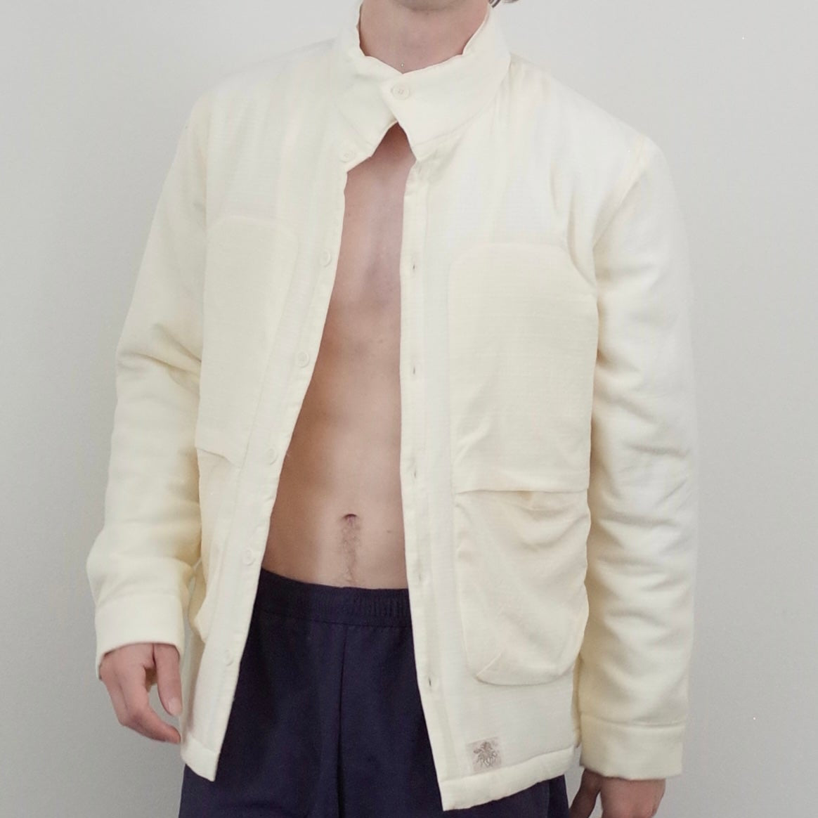 A man with curly brown hair wearing a cream-colored quilted jacket with a high collar and front pockets, styled open over a bare torso and dark navy pants, against a plain light gray background.